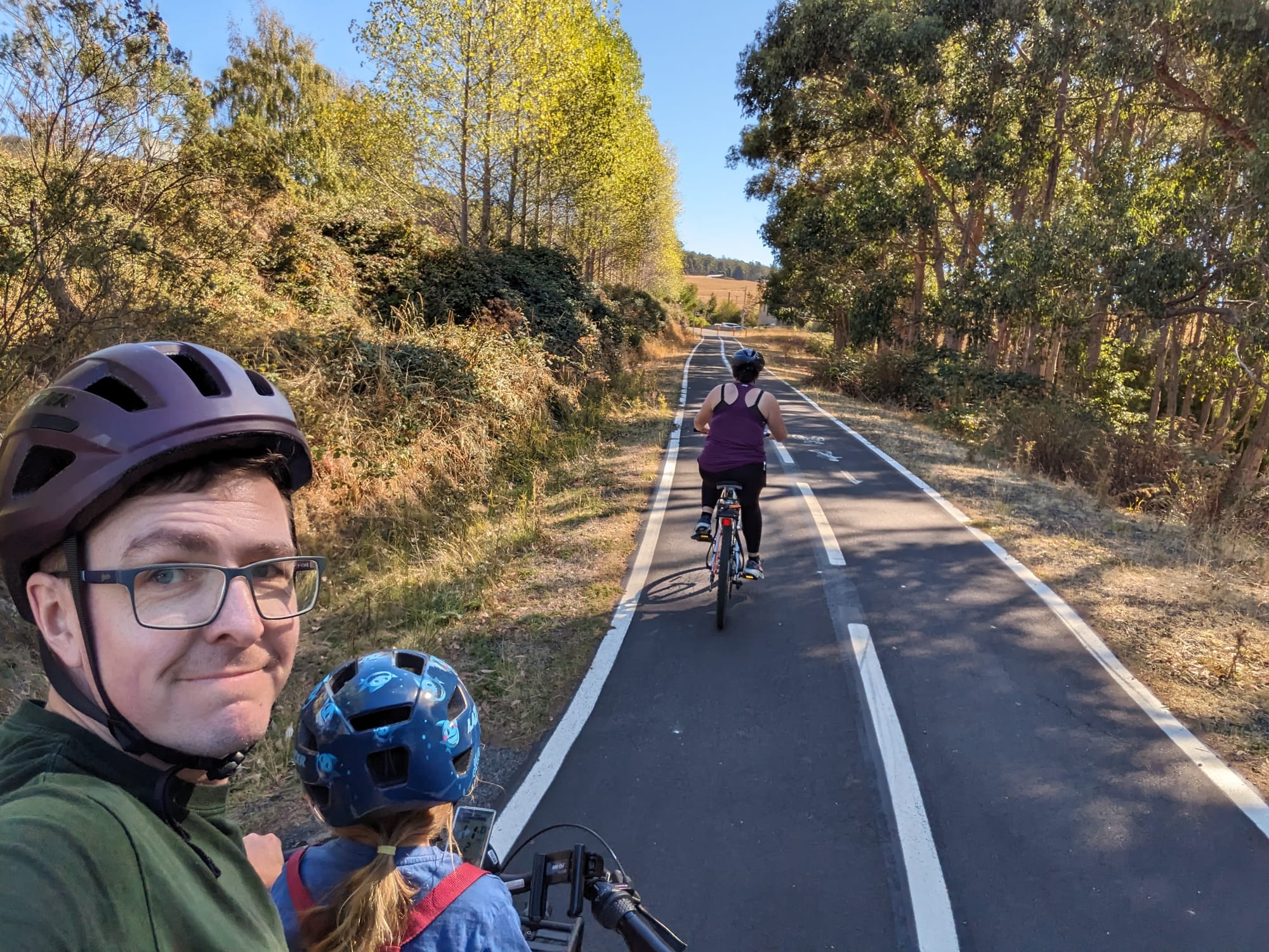 A family rides bikes on a tree-lined path. A man takes a selfie with a child in front, another rider ahead.